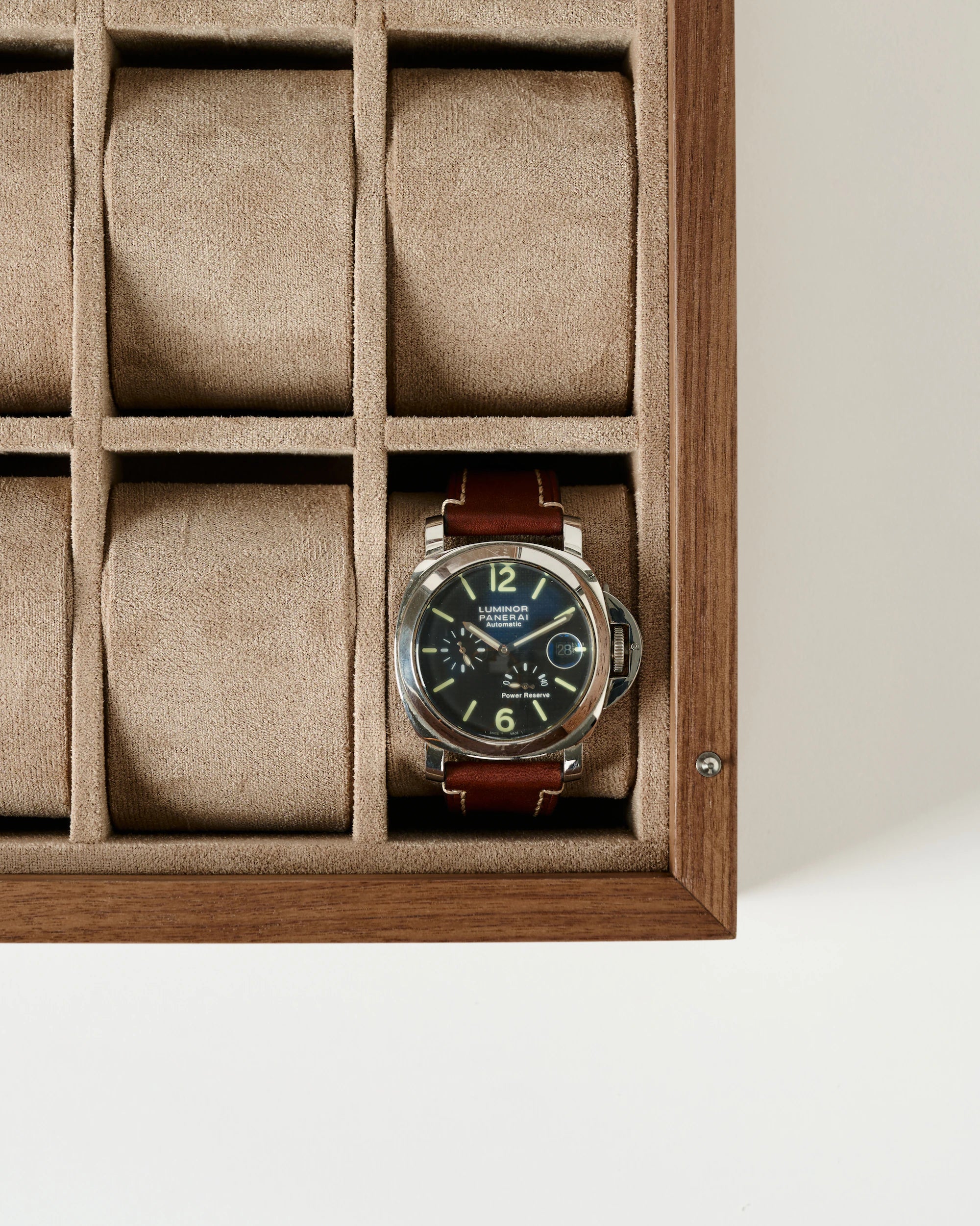 A brown leather strap wristwatch with a black dial is placed in the lower left compartment of the TAWBURY Grove 12 Slot Wooden Watch Box in Light Walnut Wood with a glass lid.