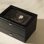 The TAWBURY Bayswater 6 Watch Jewelry Box in Black/Charcoal features a glass lid displaying several watches, with one watch face clearly visible, all set on a light-colored surface.