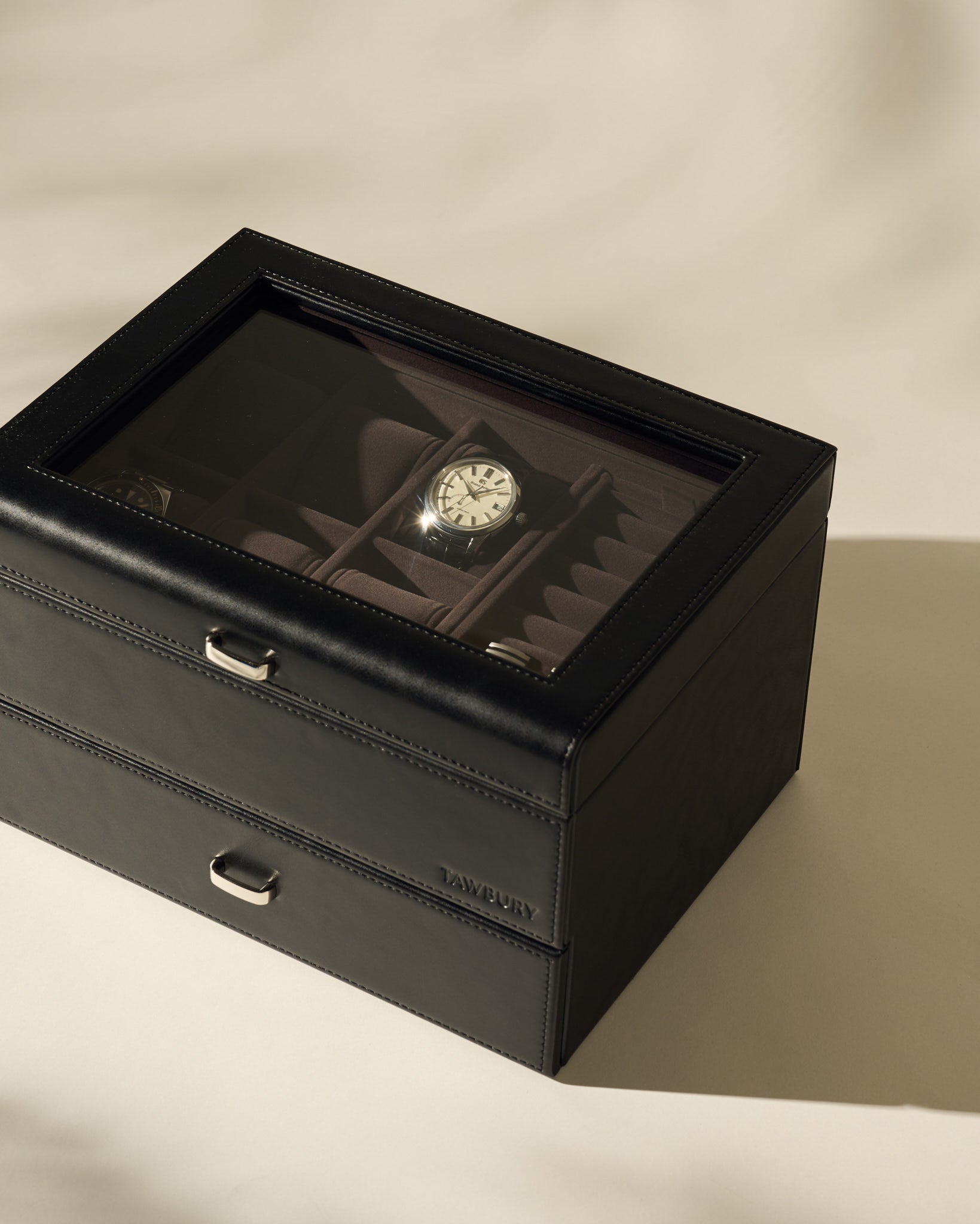 The TAWBURY Bayswater 6 Watch Jewelry Box in Black/Charcoal features a glass lid displaying several watches, with one watch face clearly visible, all set on a light-colored surface.