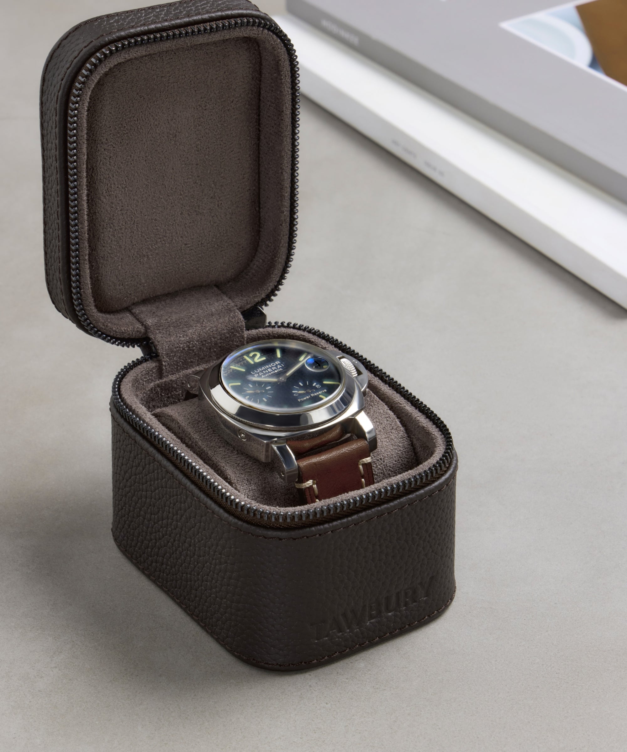 A wristwatch with a brown leather strap is displayed inside an open TAWBURY Fraser 1 Watch Travel Case - Brown (Coming Soon) on a gray surface.