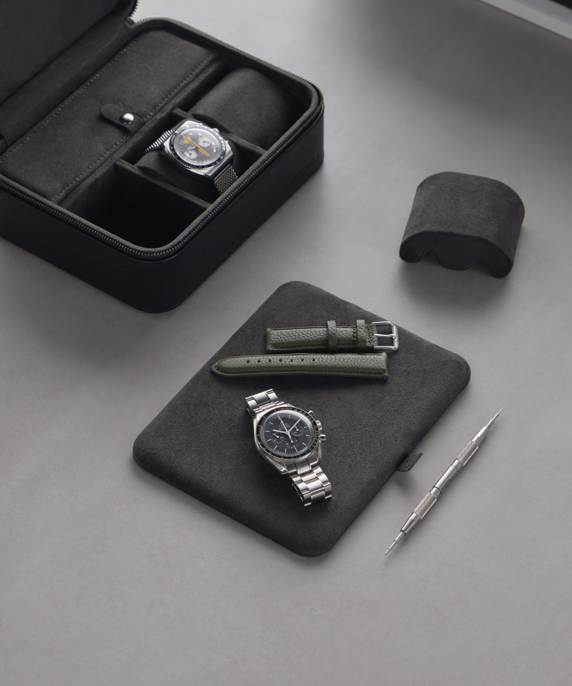 Watches and straps are elegantly displayed with a tool, as the TAWBURY Fraser 3 Watch Travel Case with Storage - Black reveals a watch inside its open lid, all set on a sleek gray surface.