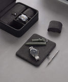 Watches and straps are elegantly displayed with a tool, as the TAWBURY Fraser 3 Watch Travel Case with Storage - Black reveals a watch inside its open lid, all set on a sleek gray surface.