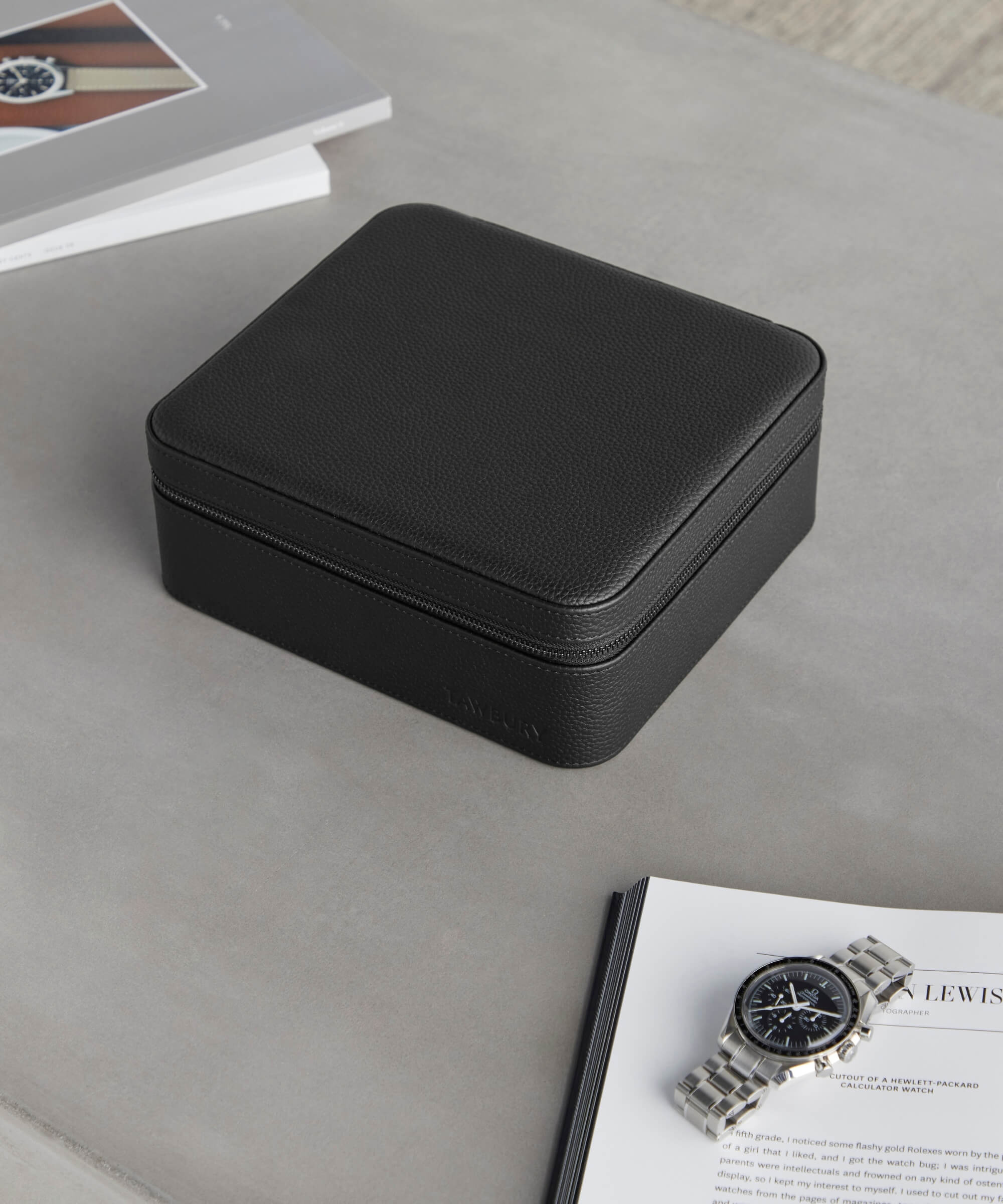 A black leather Fraser 6 Watch Travel Case - Black (Coming Soon) from the TAWBURY range sits on a table beside an open watch magazine and a wristwatch with a stainless steel band.