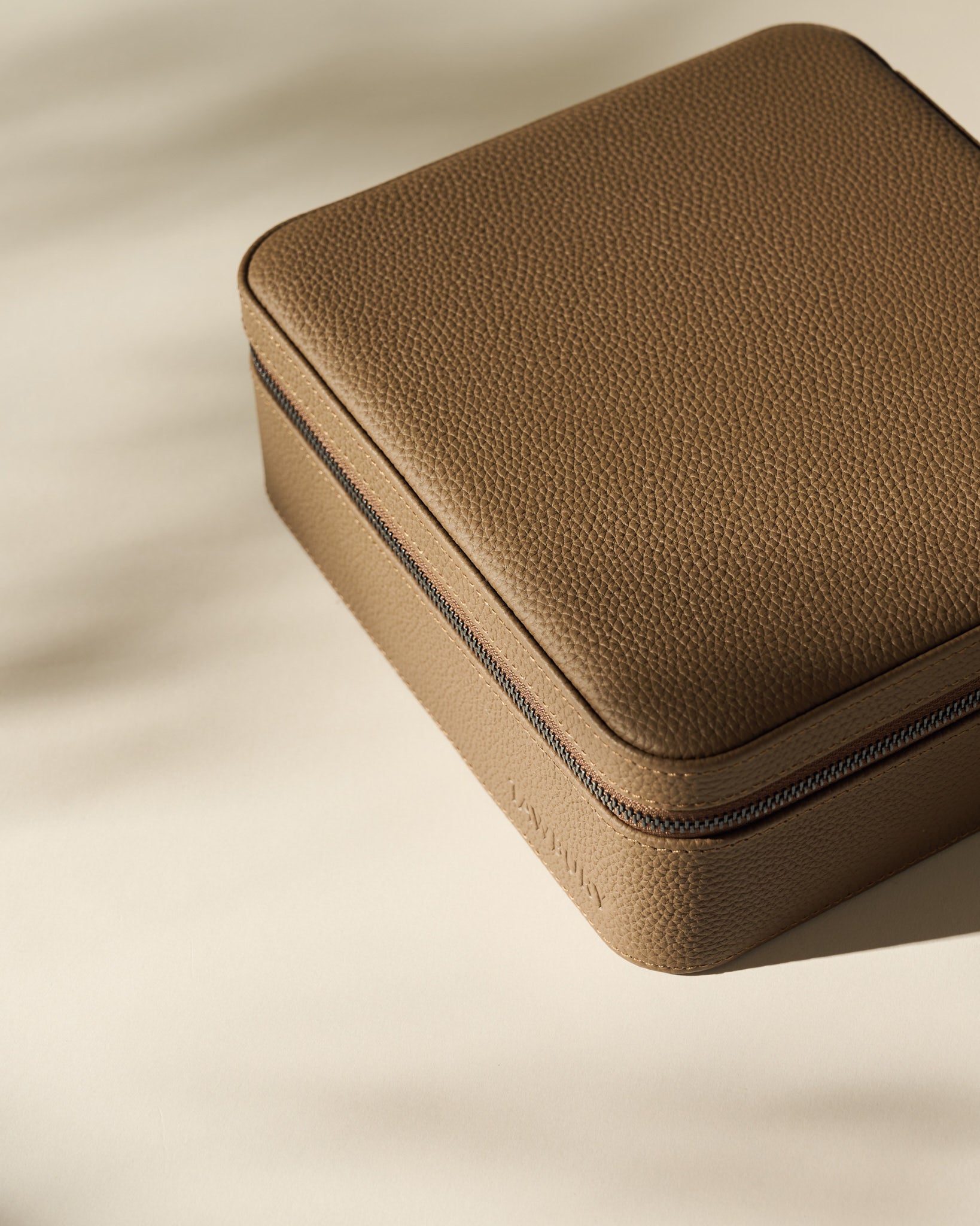 The TAWBURY Fraser 6 Watch Case in taupe features a square, textured leather design with a zipper, shown on a light surface with soft shadows.