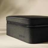 The Fraser 6 Watch Case by TAWBURY is a black textured rectangular leather box with a zippered closure, featuring "TAWBURY" embossed on the side, displayed against a neutral background.