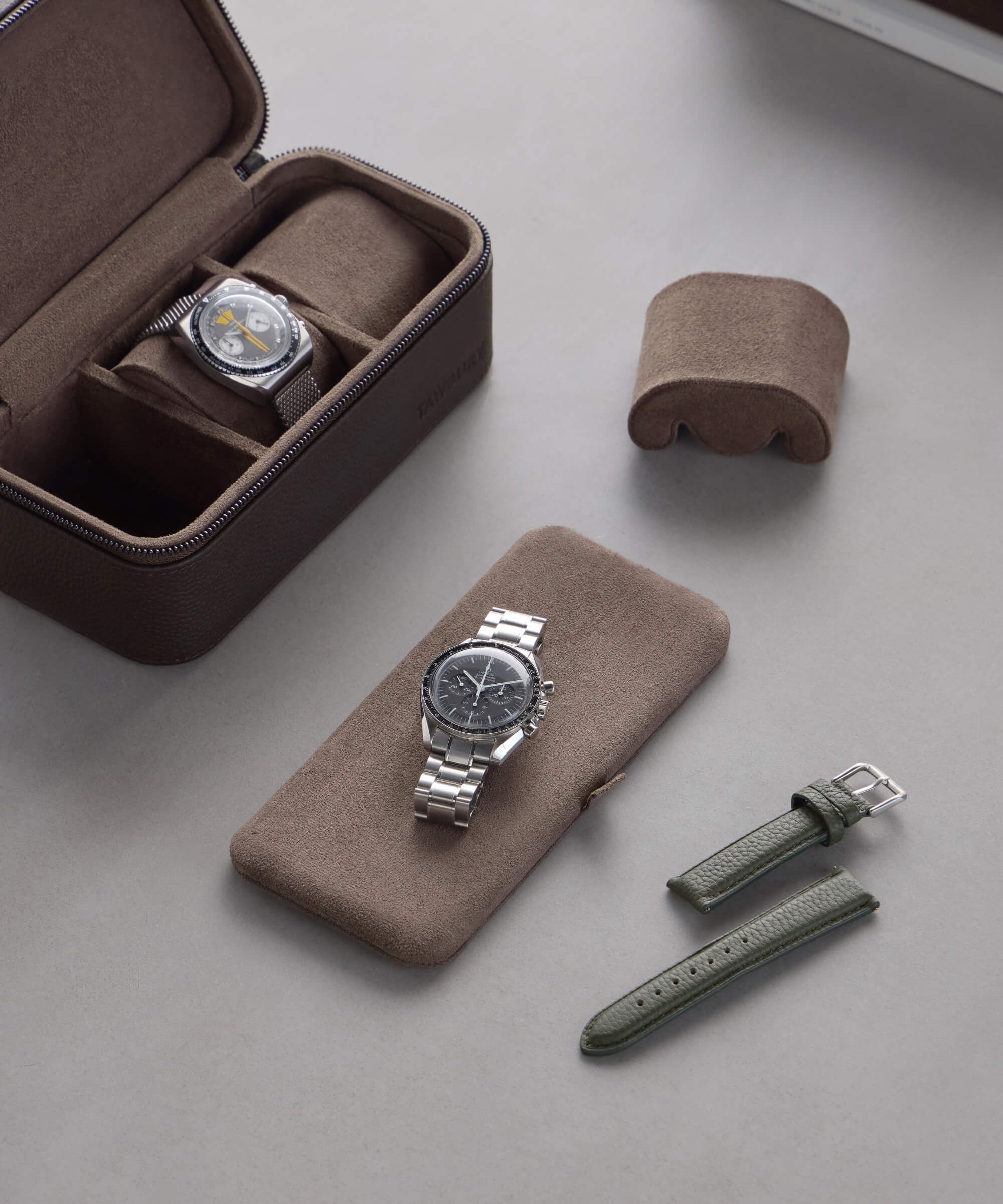 Two wristwatches are elegantly displayed: one in a Fraser 3 Watch Travel Case - Brown and the other on a cushion. Nearby, a green strap with matching buckle lies on the sleek gray surface, showcasing TAWBURY's refined style in leather watch cases.