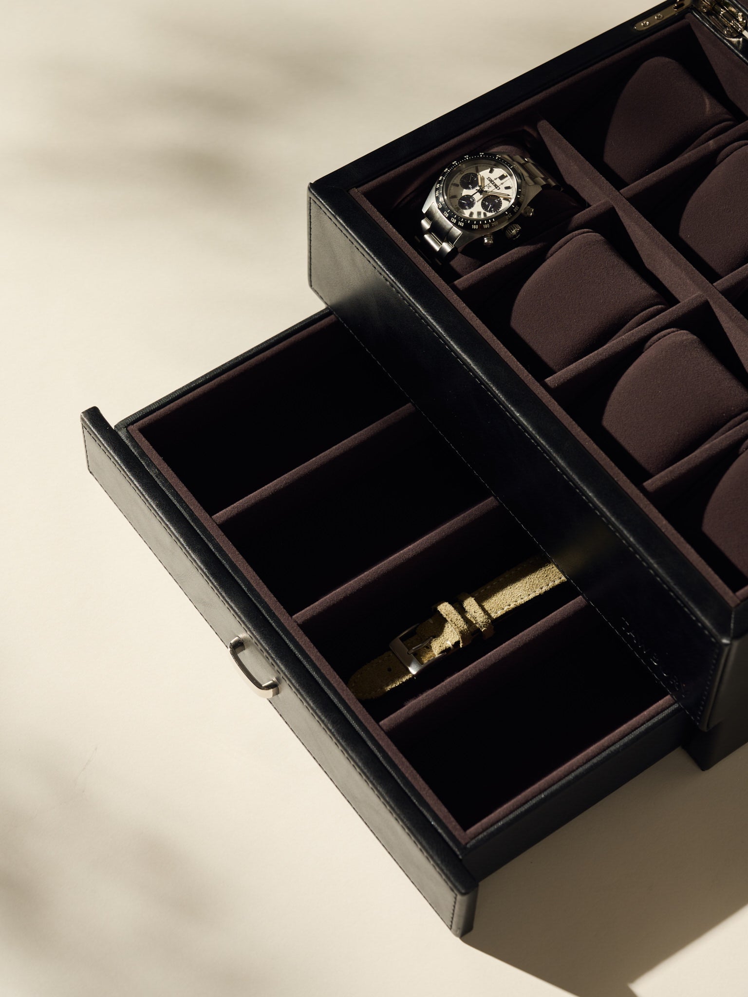 The TAWBURY Bayswater 8 Slot Watch Box with Storage in Black/Charcoal is open, elegantly displaying a watch on top, while the lower drawer stores a beige watch strap for organized storage.