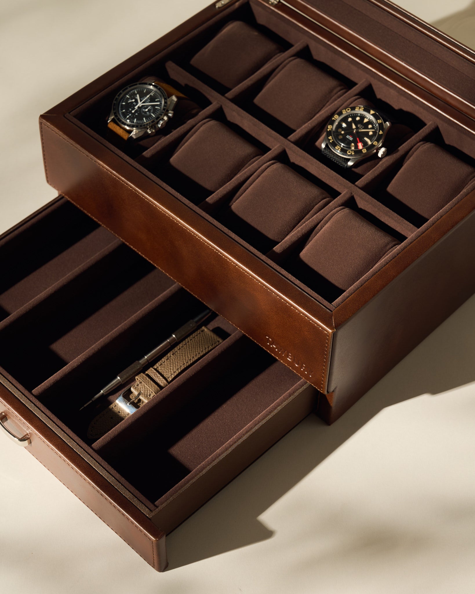 The TAWBURY Bayswater 8 Slot Watch Box with Storage in brown faux leather displays two wristwatches and a pen, with several empty compartments for organized watch storage.
