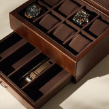The TAWBURY Bayswater 8 Slot Watch Box with Storage in brown faux leather displays two wristwatches and a pen, with several empty compartments for organized watch storage.