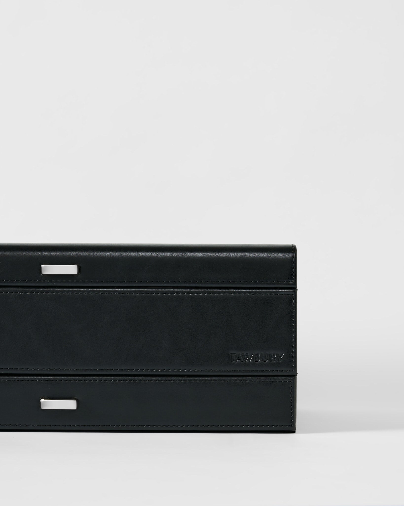 A close-up of the TAWBURY Bayswater 12 Slot Watch Box with Storage in black/charcoal, featuring silver rectangular accents and “TAWBURY” embossed on the front, set against a plain light background.