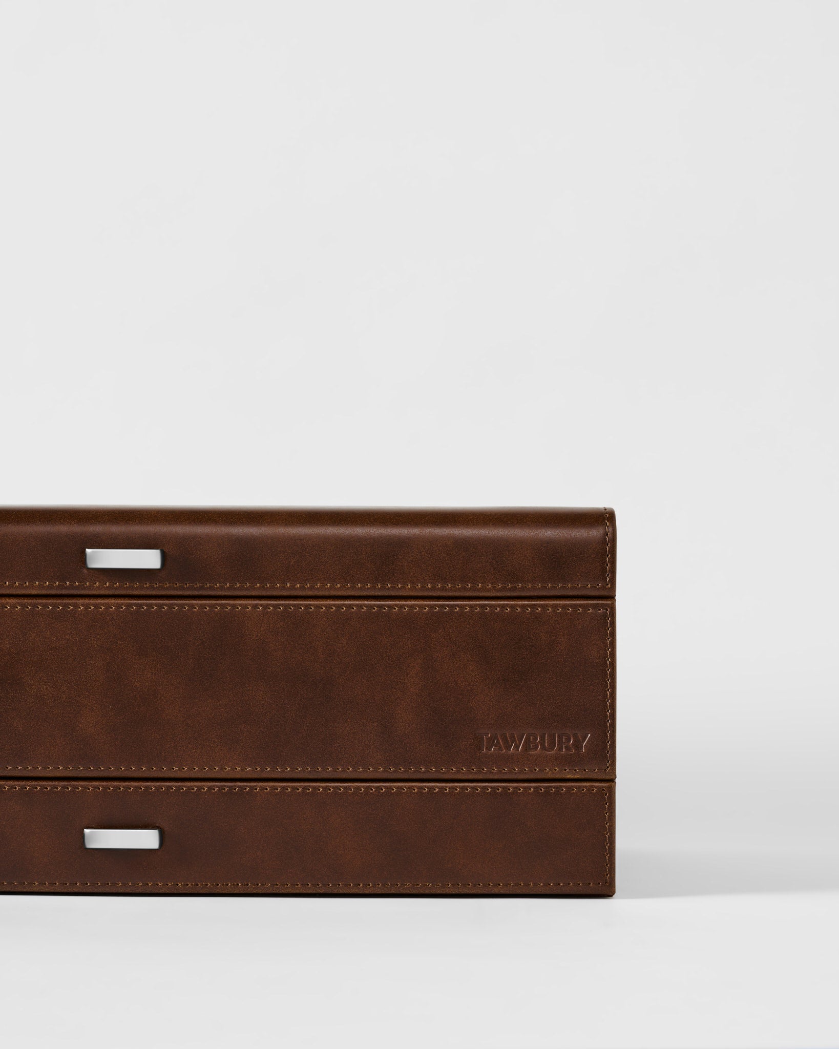 A close-up of the TAWBURY Bayswater 12 Slot Watch Box with Storage in brown, featuring silver rectangular handles and "TAWBURY" embossed on the lower right corner—ideal for elegant watch organization.