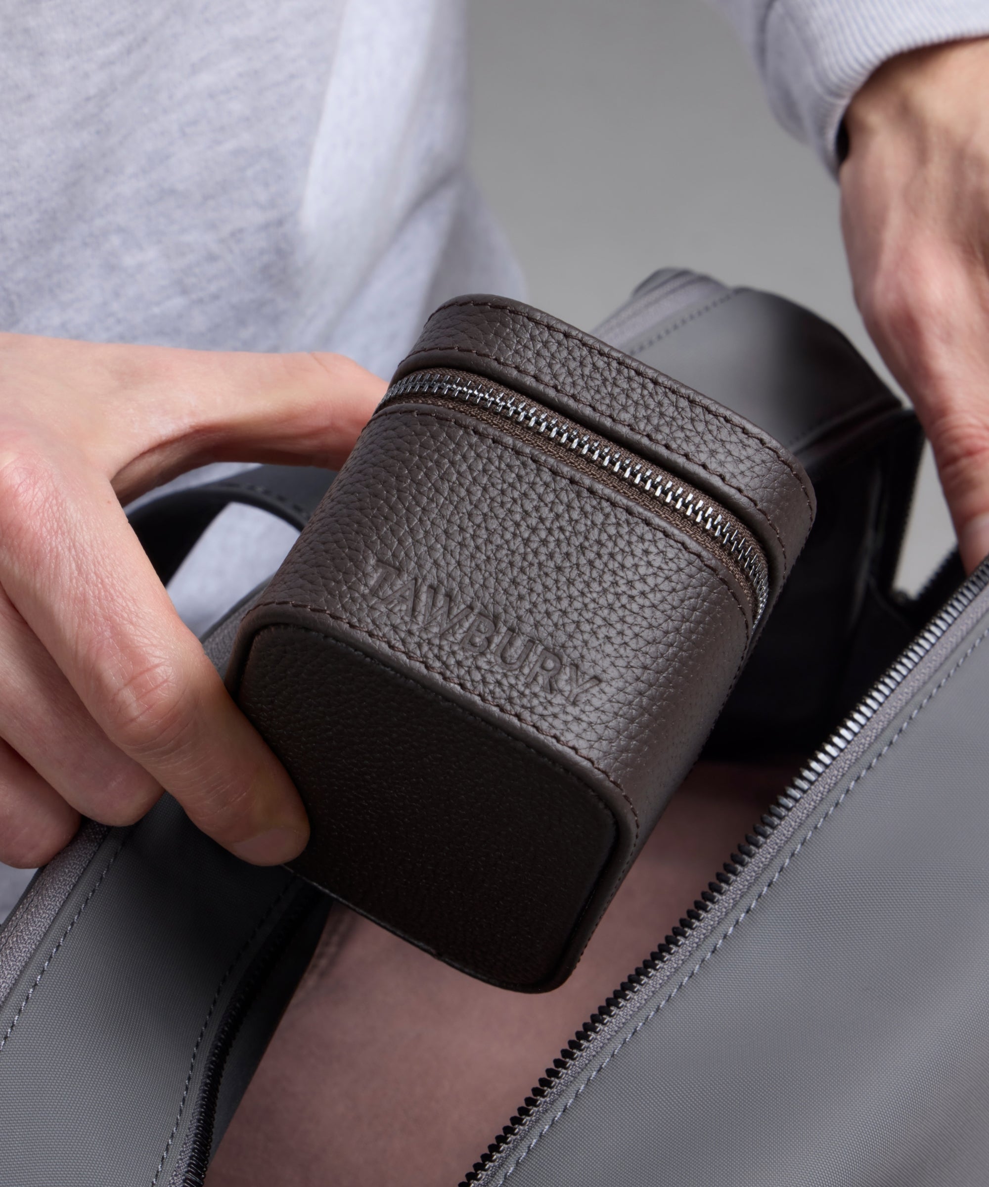 A person places the TAWBURY Fraser 1 Watch Travel Case - Brown (Coming Soon) into a gray bag, ensuring their watch is stylishly protected wherever they go.