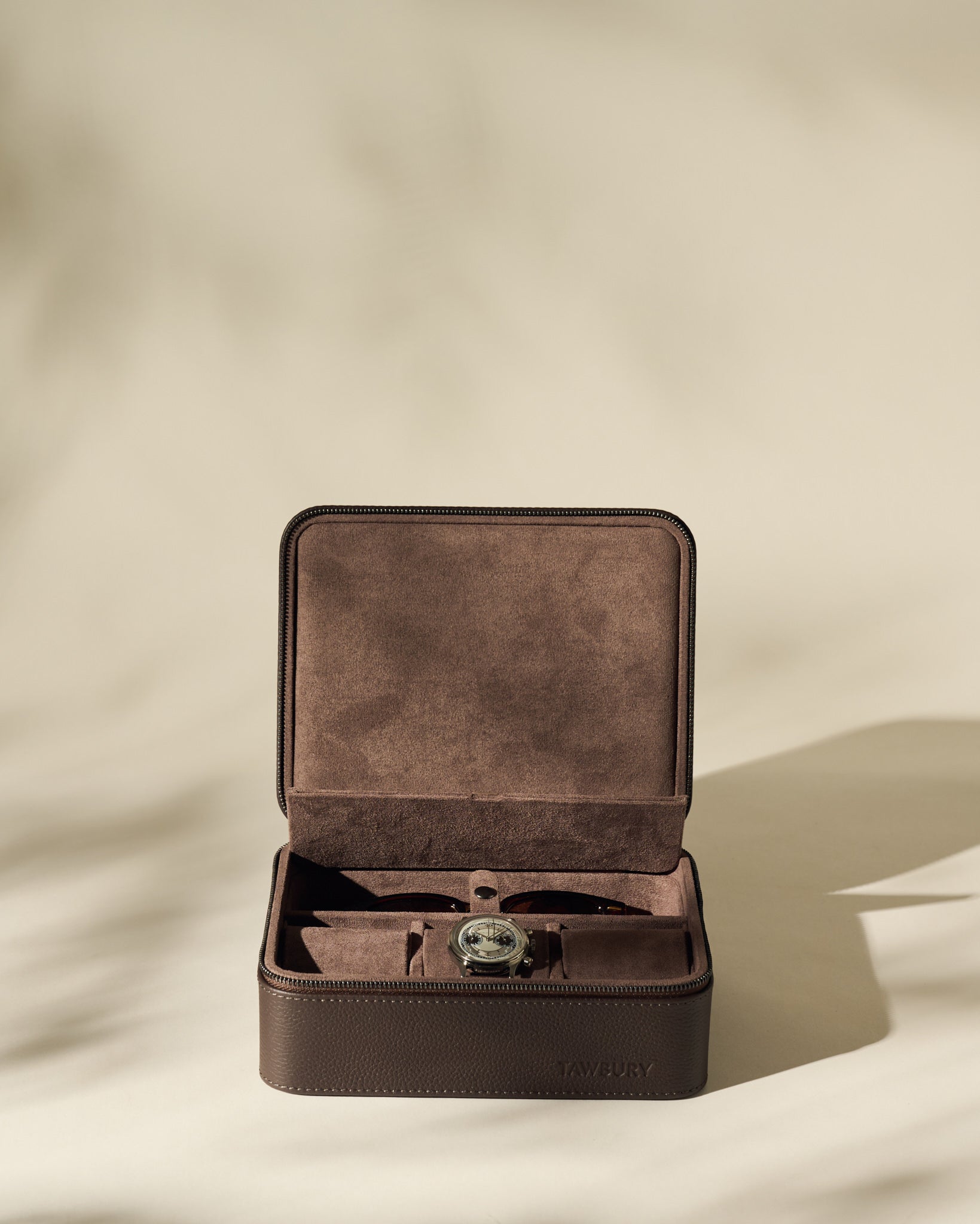 The TAWBURY Fraser 3 Watch Case with Storage in brown suede and leather sits open on a beige surface, displaying a wristwatch—ideal for storing your collection in style.