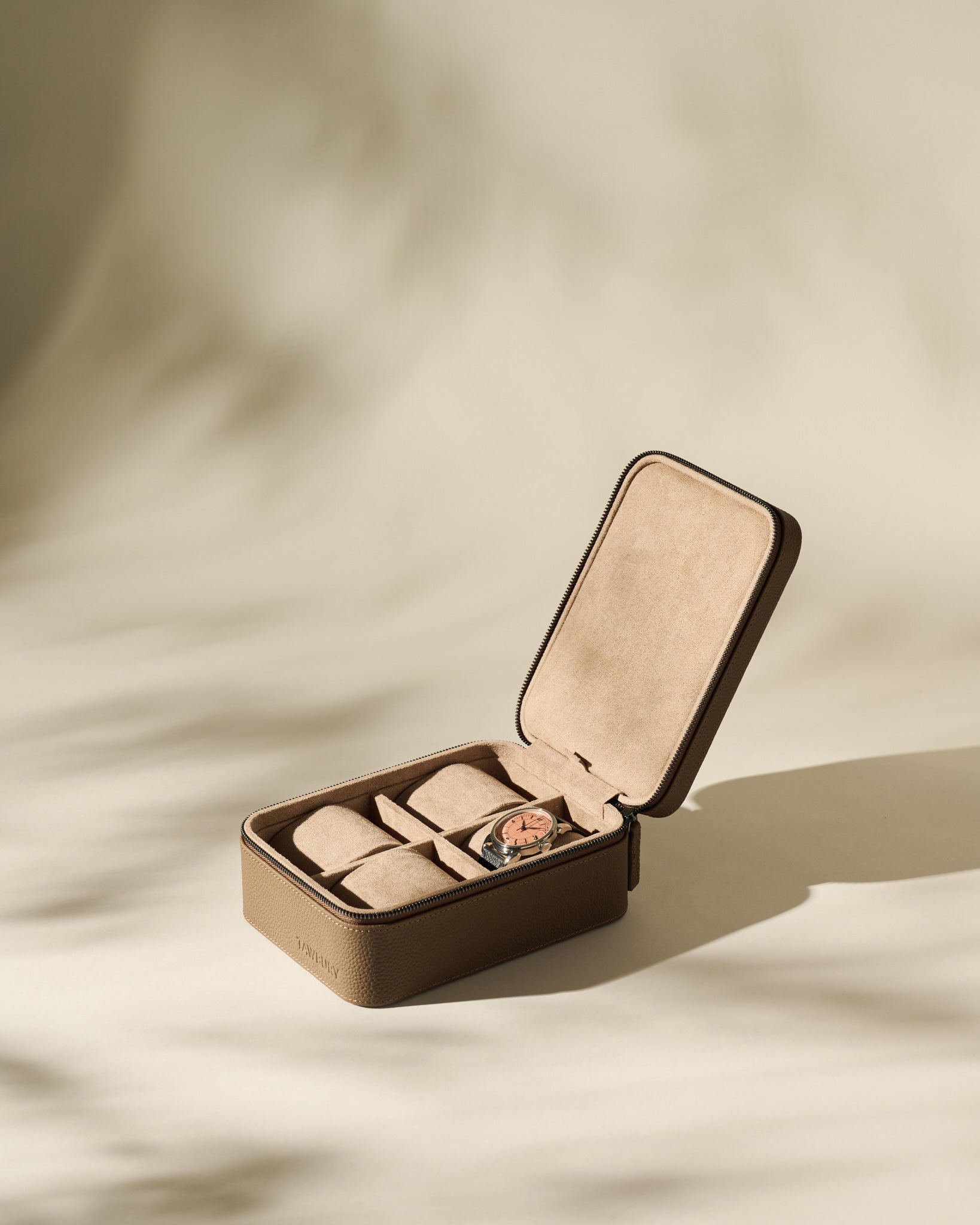 The TAWBURY Fraser 4 Watch Case in taupe is shown open on a beige surface, with multiple compartments visible and one securing a wristwatch.