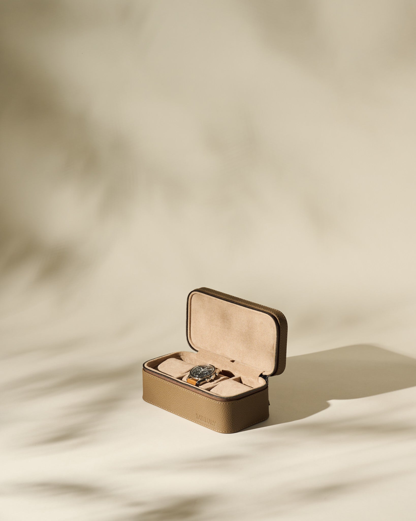 The TAWBURY Fraser 3 Watch Case in taupe, shown open with a wristwatch inside, offers stylish travel storage for your timepieces against a softly lit, neutral backdrop with gentle shadows.
