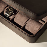 The TAWBURY Fraser 8 Watch Case - Brown, partially unzipped on a light surface, reveals its multiple compartments while storing a stainless steel wristwatch with a black dial.