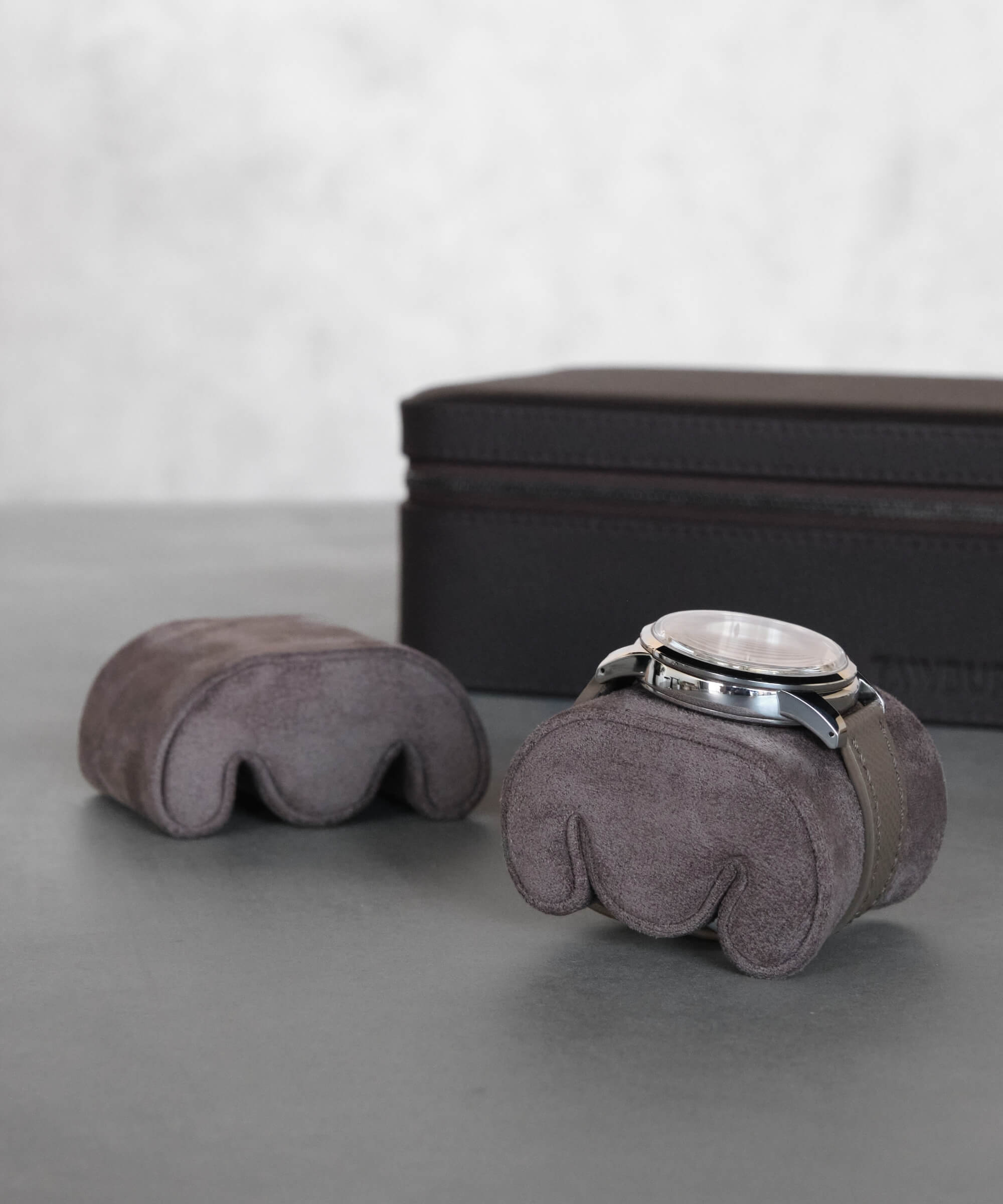 A silver wristwatch on a gray suede watch pillow is placed near a similar empty pillow, with a dark brown rectangular TAWBURY Fraser 6 Watch Travel Case - Black (Coming Soon) in the background.