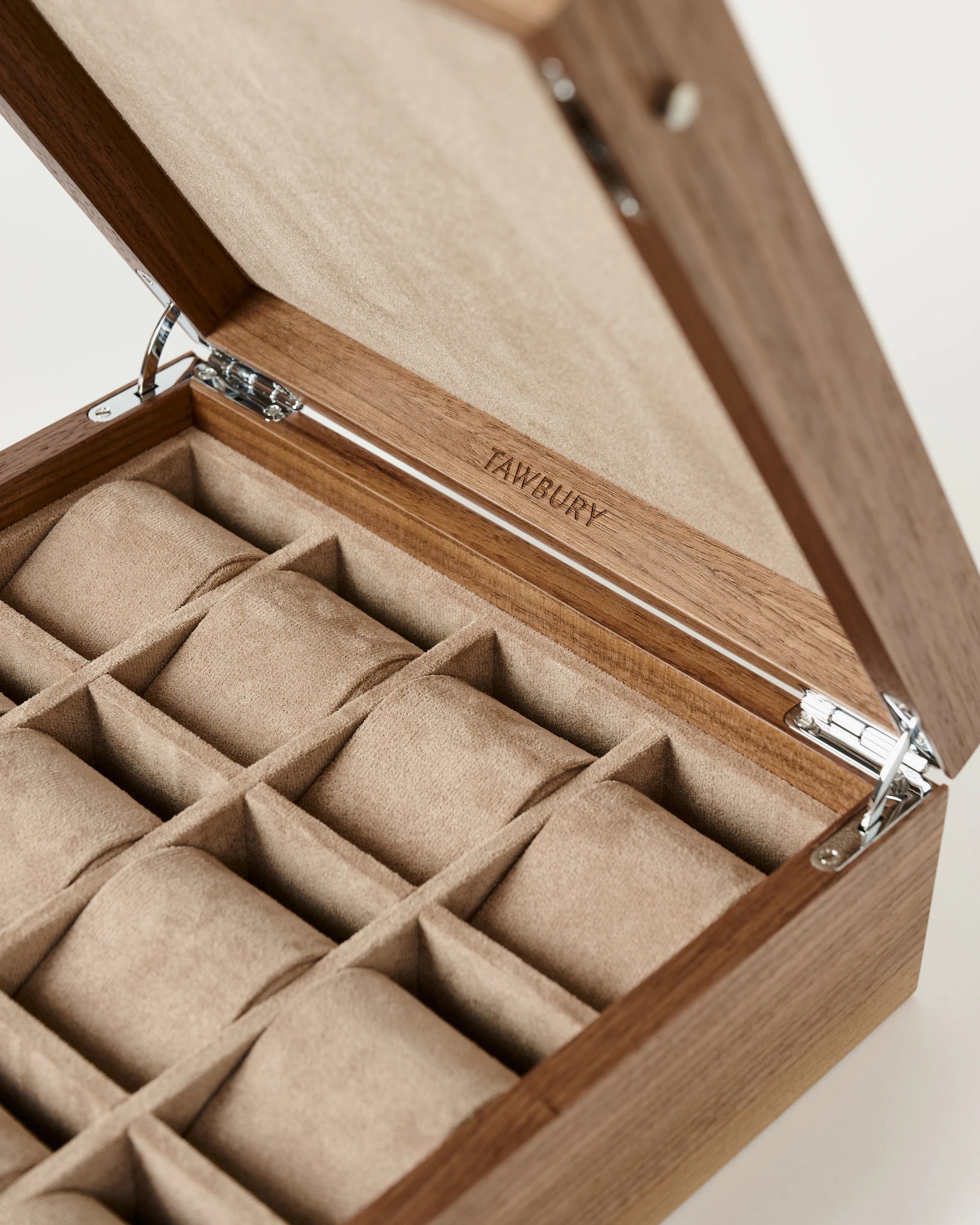 The Grove 12 Slot Wooden Watch Box by TAWBURY features a solid hinged lid, soft beige interior, and multiple compartments for watch storage. Branded with "TAWBURY" inside the lid, this elegant box is made from light walnut wood.