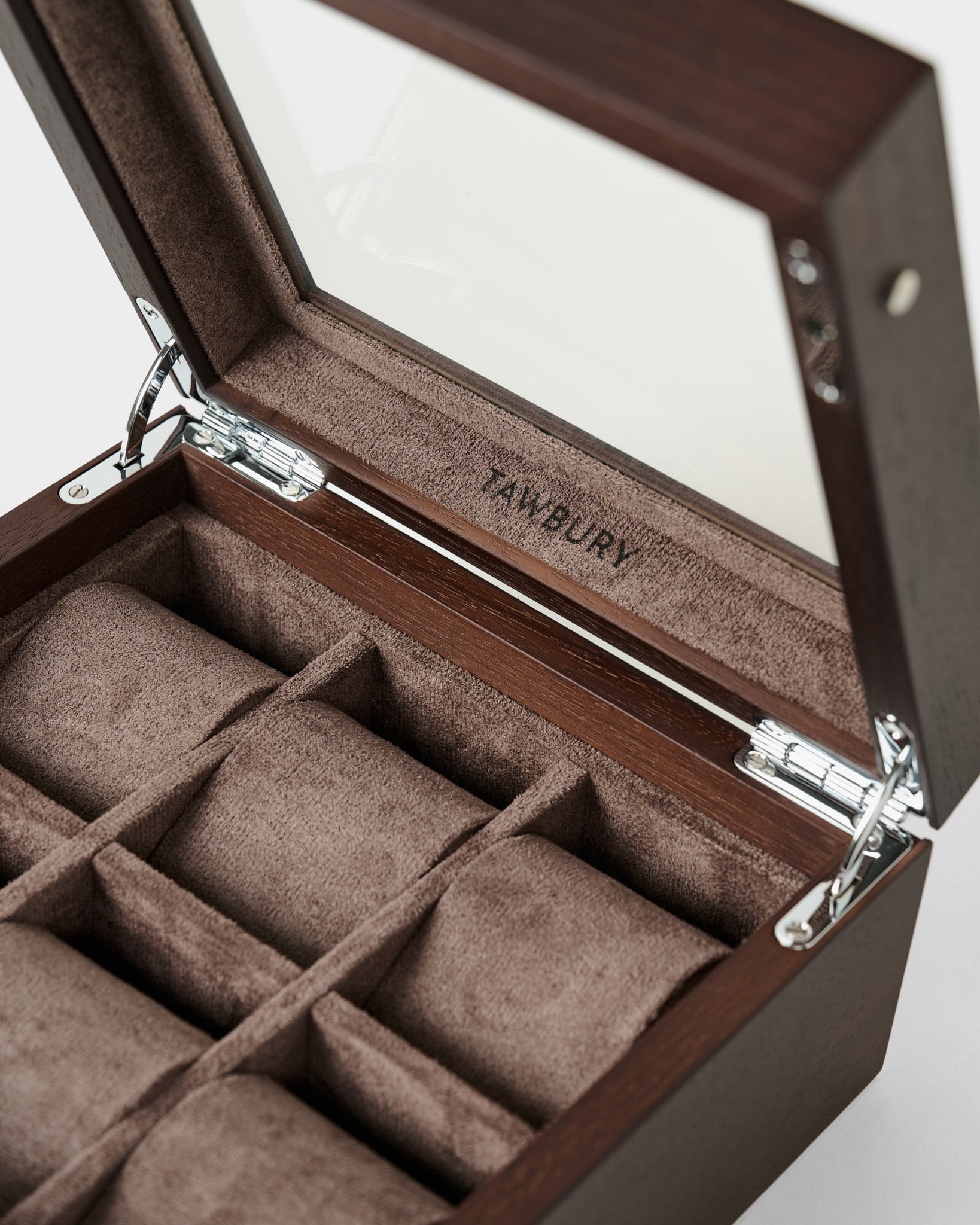 The TAWBURY Grove 6 Slot Wooden Watch Box features a dark walnut wood finish, glass lid, metal hinges, and six brown fabric-lined compartments—ideal for elegant watch storage. "TAWBURY" is printed inside.