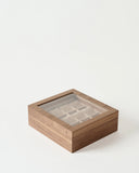 The TAWBURY Grove 12 Slot Wooden Watch Box in light walnut with a glass lid is shown on a plain white background, featuring multiple compartments for storing watches.