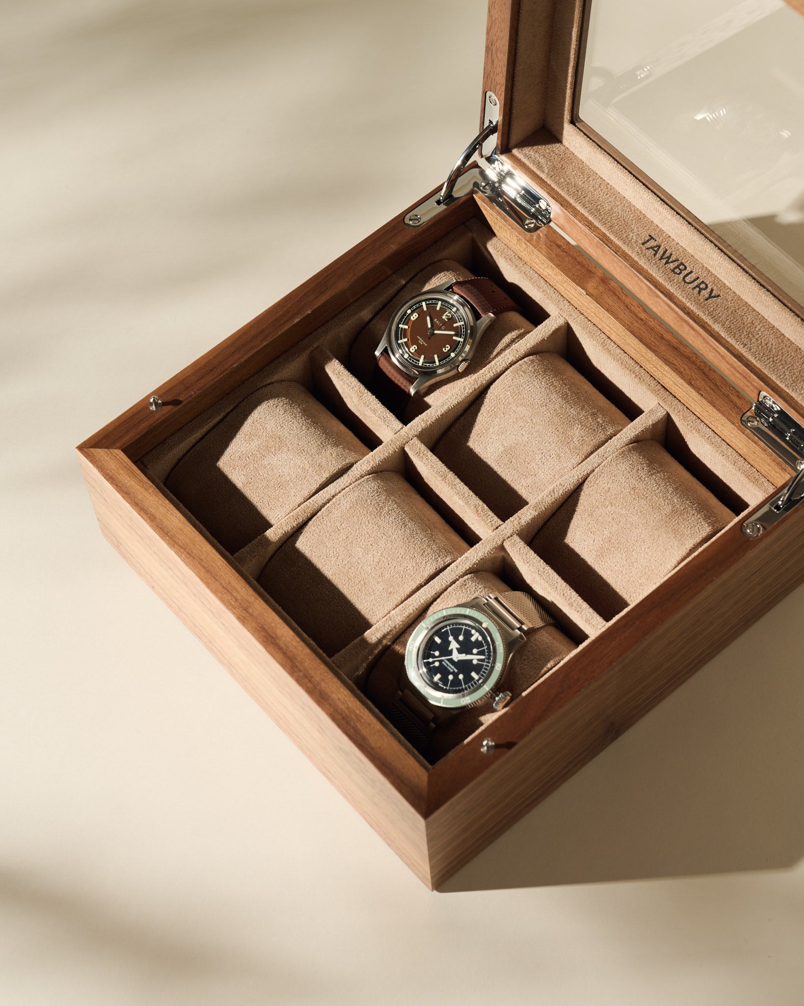 The TAWBURY Grove 6 Slot Wooden Watch Box in light walnut wood features a glass lid, two cushioned watch slots, and two empty beige suede-lined compartments—an elegant storage choice for watch enthusiasts.