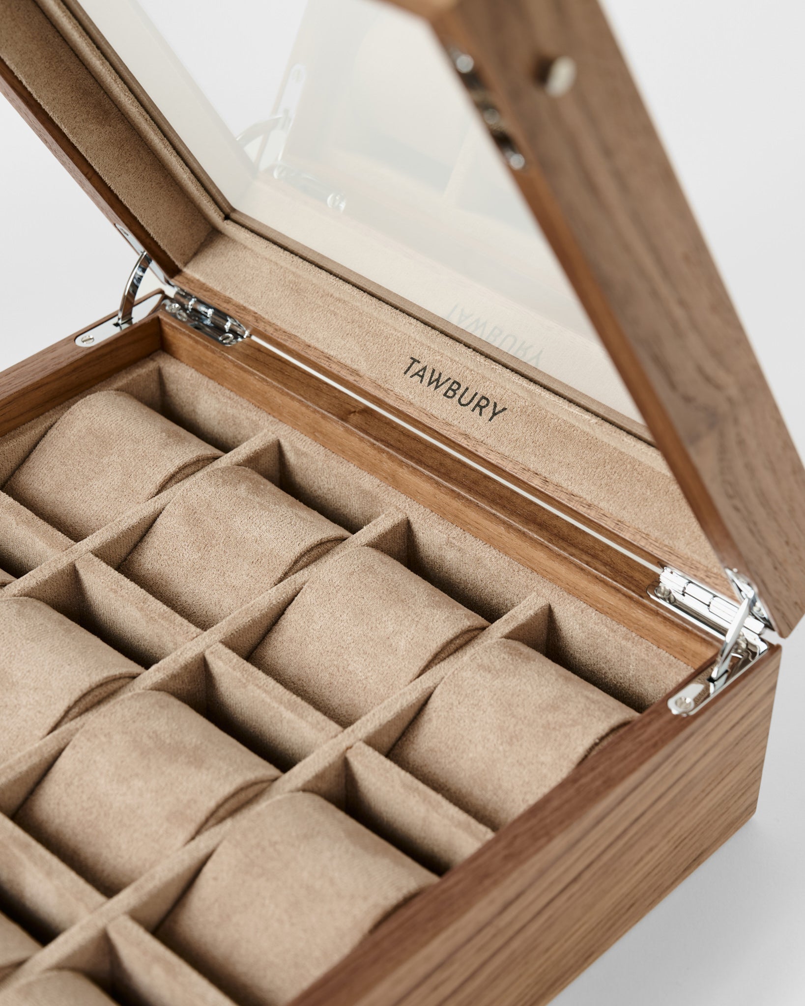 The TAWBURY Grove 12 Slot Wooden Watch Box features a light walnut finish, glass lid, beige suede interior, and multiple compartments for storing watches, with the TAWBURY logo inside the lid.