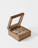 The TAWBURY Grove 6 Slot Wooden Watch Box in light walnut features a glass lid, metal hinges, and six cushioned compartments—ideal for watch collectors—shown against a plain white background.