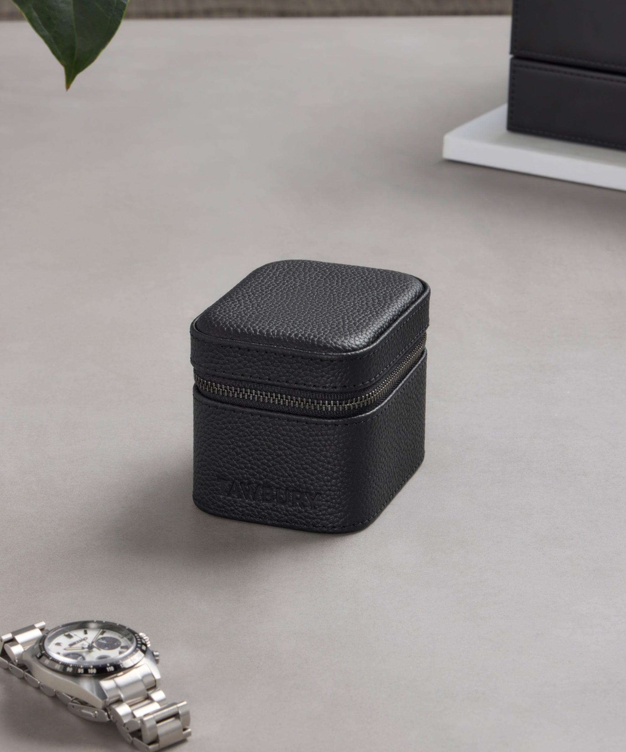 The TAWBURY Fraser 1 Watch Travel Case - Black (Coming Soon) is a black textured leather zippered case on a gray surface near a silver watch, providing stylish and reliable protection for your timepiece while traveling.