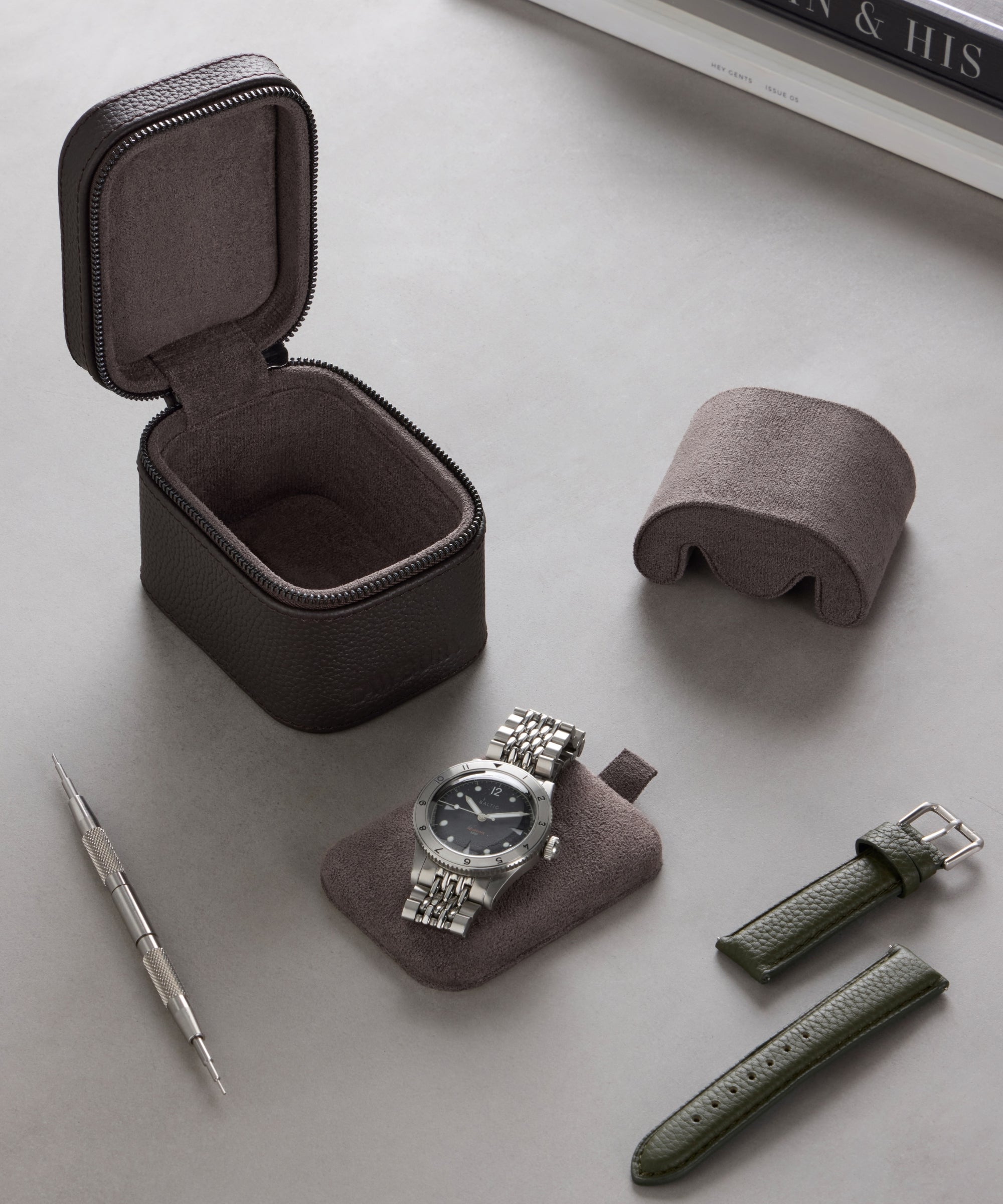 A silver wristwatch, tool, and two green straps are displayed with the TAWBURY Fraser 1 Watch Travel Case - Brown (Coming Soon) on a gray surface beside stacked books.