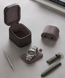 A silver wristwatch, tool, and two green straps are displayed with the TAWBURY Fraser 1 Watch Travel Case - Brown (Coming Soon) on a gray surface beside stacked books.