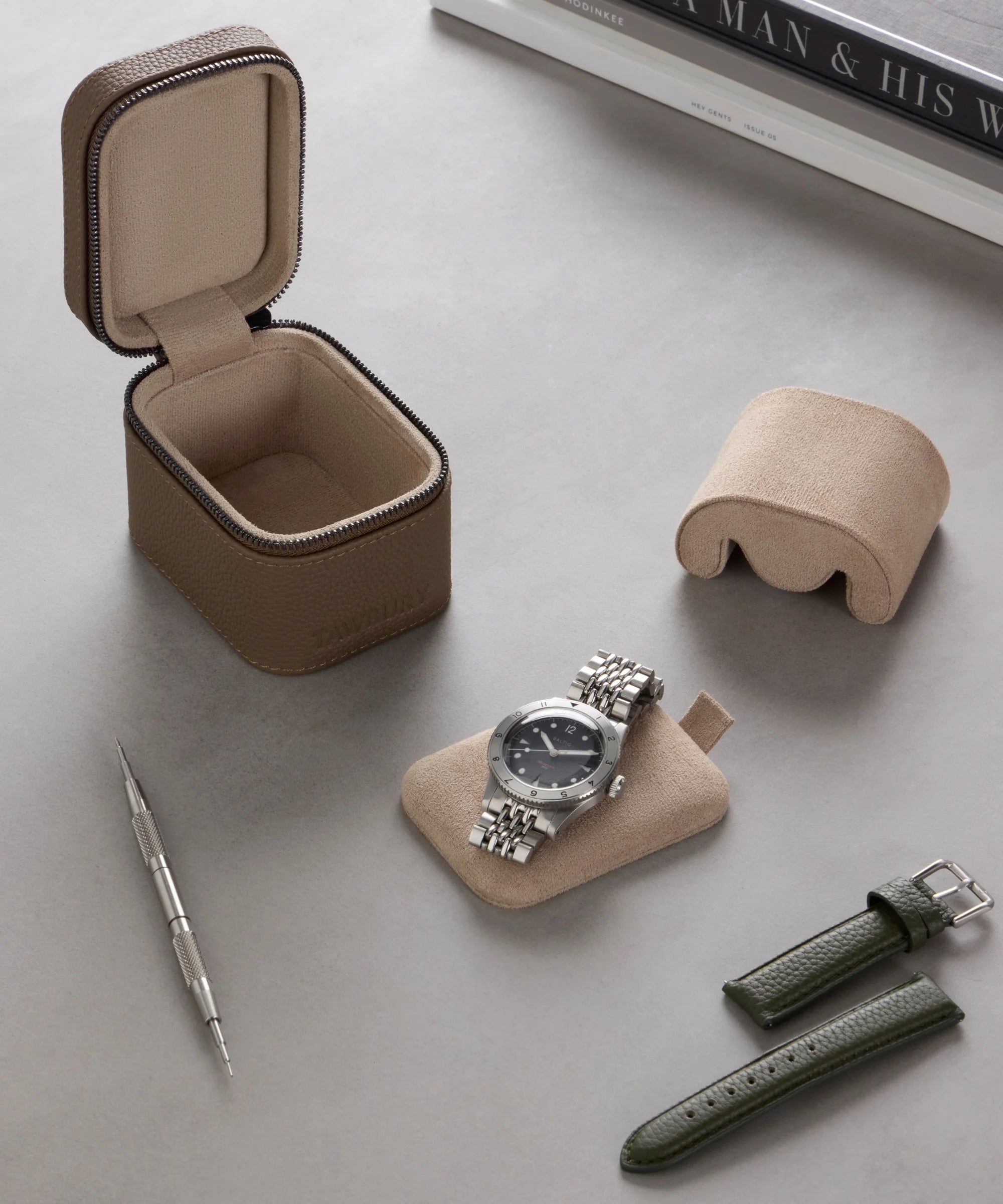 A wristwatch with a metal band rests on a padded holder beside the TAWBURY Fraser 1 Watch Travel Case - Taupe (Coming Soon), green leather straps, a tool, and a cushion, all arranged on a gray surface.