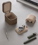 A wristwatch with a metal band rests on a padded holder beside the TAWBURY Fraser 1 Watch Travel Case - Taupe (Coming Soon), green leather straps, a tool, and a cushion, all arranged on a gray surface.